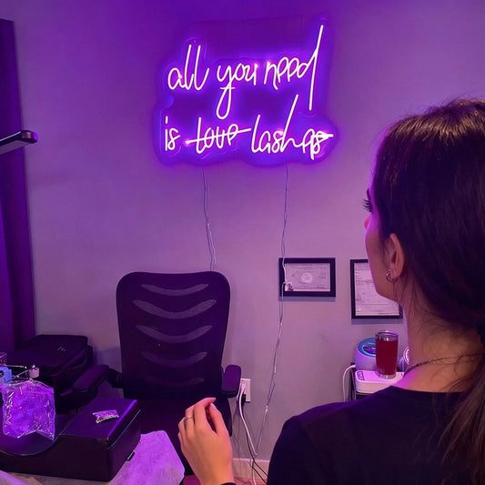 Woman standing in front of a chair with a neon sign on the wall reading 'all you need is love lashes' in a room.