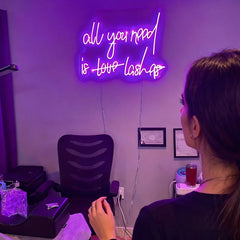 Woman standing in front of a chair with a neon sign on the wall reading 'all you need is love lashes' in a room.