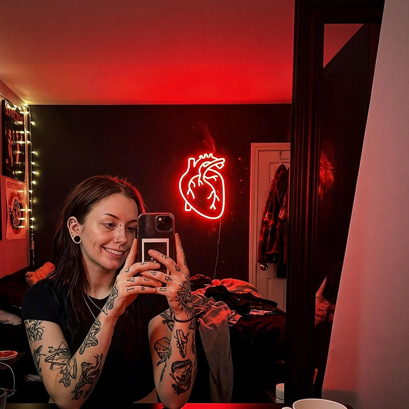 Person with tattoos using a phone in a room with a heart-shaped neon light.