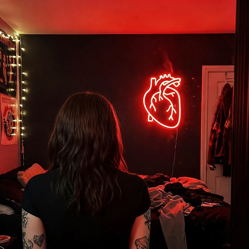 Person with tattoos sitting in a room with a red neon heart sign on the wall.
