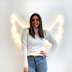 Woman wearing a white long-sleeve top and blue jeans standing in front of a wall with angel wings made of neon lights.