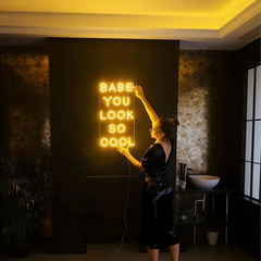 Woman in a dark room holding a neon sign that says 'Babe you look so cool'.