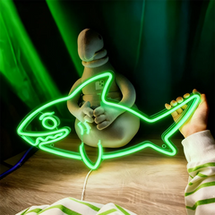 Neon green fish-shaped light being held by a person on a wooden surface with a dark green curtain background.