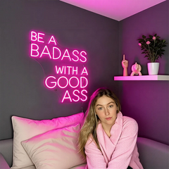 Woman sitting on a couch with a neon sign on the wall that reads 'Be a Badass with a Good Ass'.