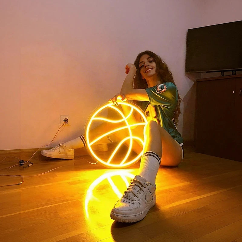 Person sitting on a wooden floor with a glowing lemon yellow color basketball neon sign.