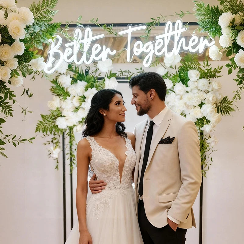 Couple in wedding attire standing in front of a floral arch with 'Better Together' sign.