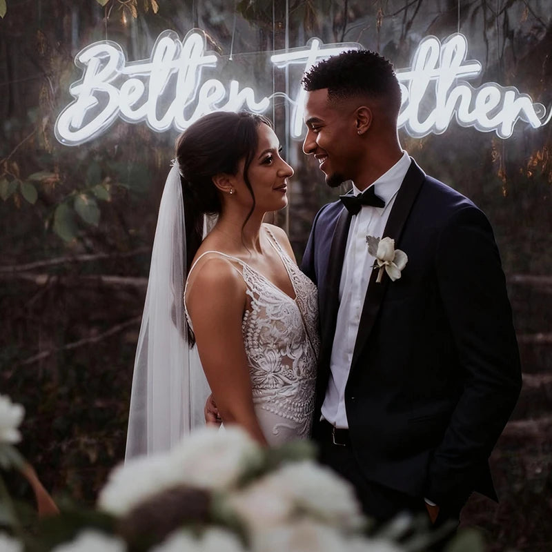 Couple in wedding attire standing in front of a neon sign with the words 'Better Together' in a forest setting.