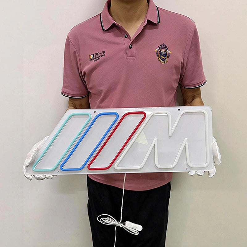 A person wearing a pink polo shirt and black pants holds a white illuminated BMW M logo sign with gloves. The sign features the iconic M logo with red, blue, and light green illuminated accents.