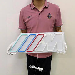 A person wearing a pink polo shirt and black pants holds a white illuminated BMW M logo sign with gloves. The sign features the iconic M logo with red, blue, and light green illuminated accents.