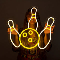 A smilling woman holding a neon sign shaped like bowling with lemon yellow and white color in a dark background