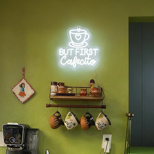 A glowing white neon-style wall sign reading “BUT FIRST Cafecito” with a coffee cup icon, mounted on a green wall above a shelf and hanging mugs in a cafe-like interior.