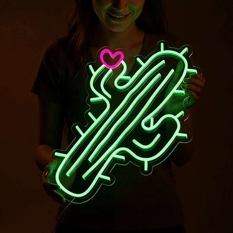 A person holds a neon light shaped like a green cactus with a small pink heart at the top. The neon glows brightly against a dark background, partially illuminating the person’s face and torso.