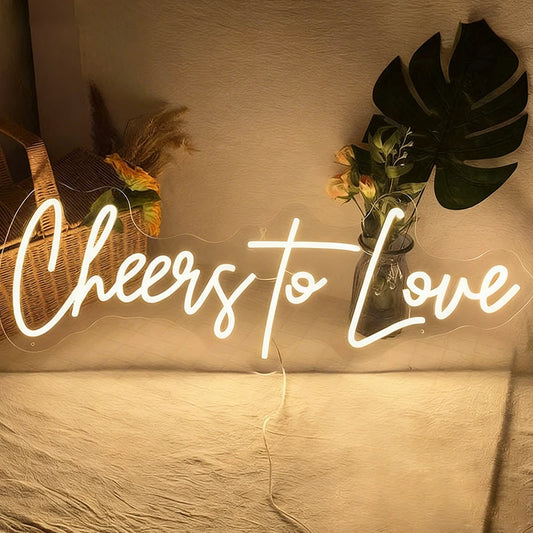 Neon sign with 'Cheers to Love' in a cozy indoor setting with plants and a basket.