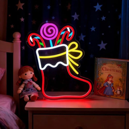 A glowing Christmas stocking LED neon sign shaped like a stocking with candy canes and a yellow bow, displayed on a bedside table next to a doll and a children's Christmas book in a bedroom setting.