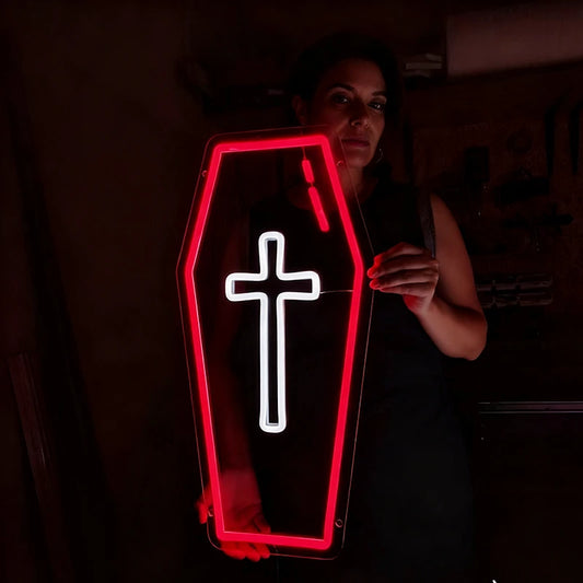 A coffin-shaped LED neon sign with a glowing red border and a white cross in the center, held by a woman in a dark room.