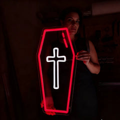 A coffin-shaped LED neon sign with a glowing red border and a white cross in the center, held by a woman in a dark room.