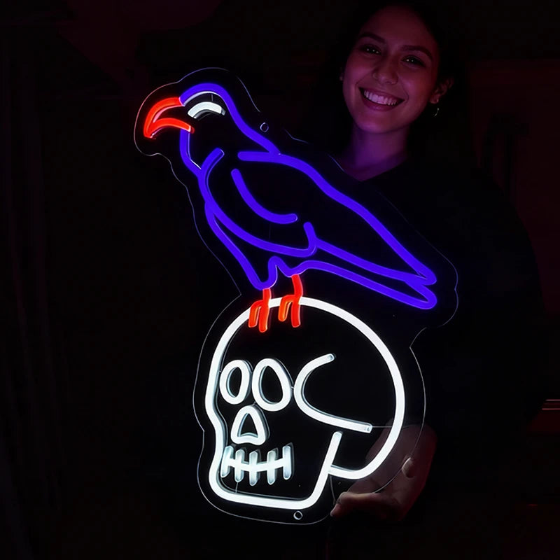 A smiling woman holds a glowing LED neon sign shaped like a white skull with a purple raven perched on top, featuring red accents on the beak and legs, set against a dark background.