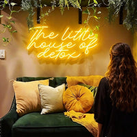 A warm lemon yellow neon sign reading 'The little house of detox' mounted on a beige wall above a green sofa with yellow and cream pillows. A woman with long wavy hair sits in the foreground, facing the sign.