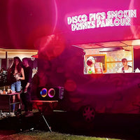 A food truck or bar cart at night with bright hot pink neon signage reading 'DISCO PIGS SMOKIN' DRINKS PARLOUR.' Two people stand beside a small table on the left, and a person is visible inside the serving window on the right.