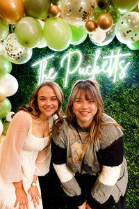 Two women posing in front of a neon sign with balloons and greenery