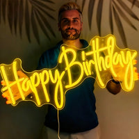 A man holding a glowing lemon yellow neon sign that reads 'Happy Birthday' against a dark background with palm leaf shadows.