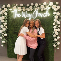 Three young women hugging in front of a decorative green wall with white flowers and a glowing neon sign that reads 'The Way Church'.
