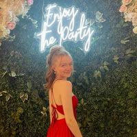 A young woman in a red dress posing in front of a green leaf wall with a glowing 'Enjoy the Party' sign and floral decorations.
