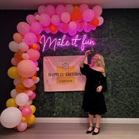 A woman in a black off-the-shoulder dress stands in front of a decorated party backdrop with pink, orange, and white balloons, a neon sign reading 'Make it fun,' and a hot pink neon sign that says 'Happy 21st Birthday Caroline.'.