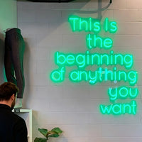 A bright green neon sign on a white brick wall reads 'This is the beginning of anything you want', with a mannequin wearing dark leggings on the left and a small plant at the bottom.