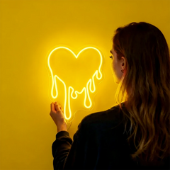 Person interacting with a neon heart-shaped light on a yellow wall
