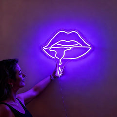 Woman holding a neon purple sign shaped like lips against a wall.