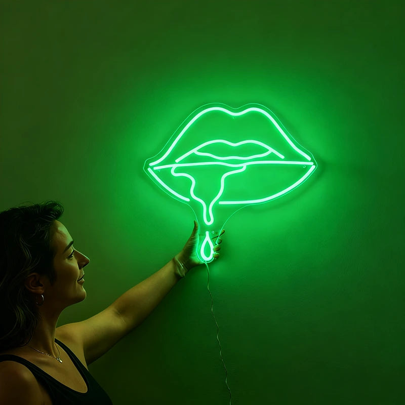 Woman holding a neon green sign shaped like lips against a wall.