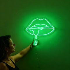 Woman holding a neon green sign shaped like lips against a wall.