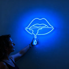 Woman holding a neon blue sign shaped like lips against a wall.