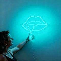 Woman holding a neon ice blue sign shaped like lips against a wall.