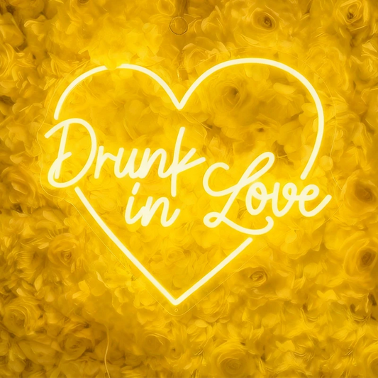 Neon lemon yellow sign with 'Drunk in Love' text in the shape of a heart on a floral background