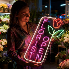 Woman holding a colorful neon sign read 'FLORIST OPEN' in a flower shop.