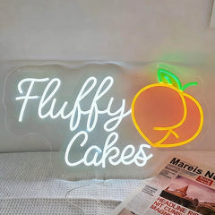 Neon sign with 'Fluffy Cakes' and a peach graphic on a white surface.