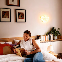 Woman reading a book on a bed in a cozy bedroom with warm white keith haring flying man neon sign on the wall.