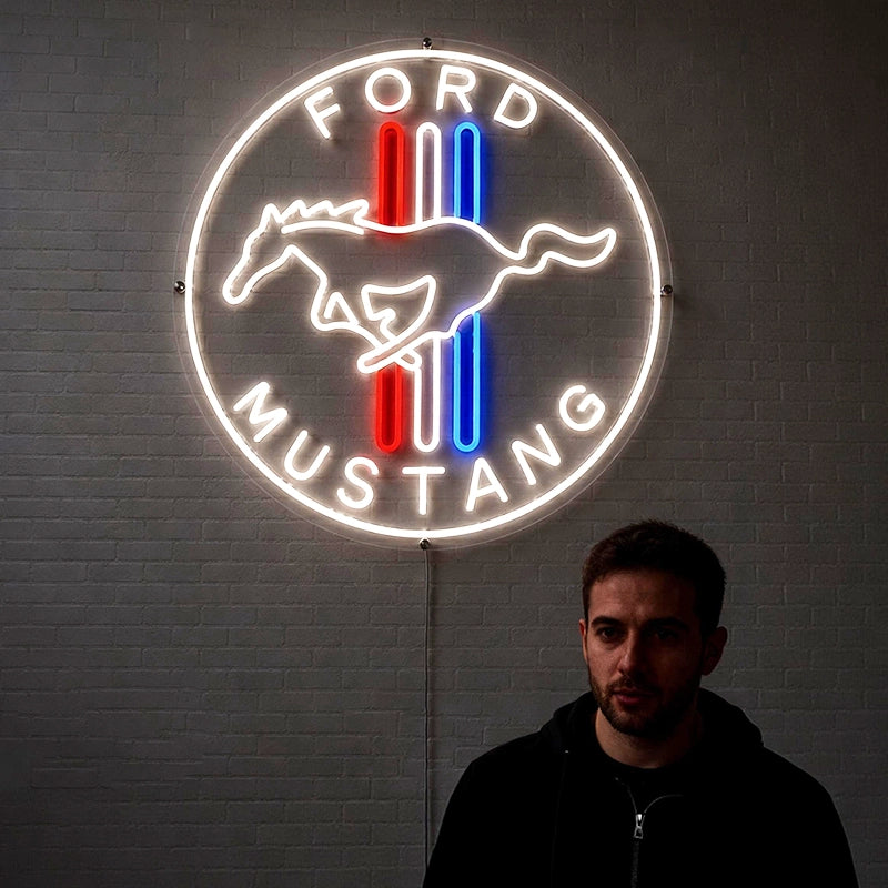 A glowing Ford Mustang neon sign mounted on a gray brick wall, with a man standing in front of it wearing a black hoodie. The sign features a white circular border, the words 'FORD MUSTANG', a white horse silhouette, and red and blue vertical stripes.