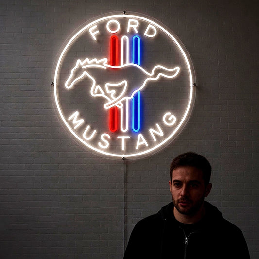 A glowing Ford Mustang neon sign mounted on a gray brick wall, with a man standing in front of it wearing a black hoodie. The sign features a white circular border, the words 'FORD MUSTANG', a white horse silhouette, and red and blue vertical stripes.