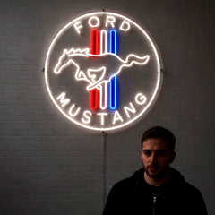 A glowing Ford Mustang neon sign mounted on a gray brick wall, with a man standing in front of it wearing a black hoodie. The sign features a white circular border, the words 'FORD MUSTANG', a white horse silhouette, and red and blue vertical stripes.