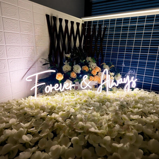 Neon sign reading 'Forever & Always' surrounded by flowers on a decorative stand.