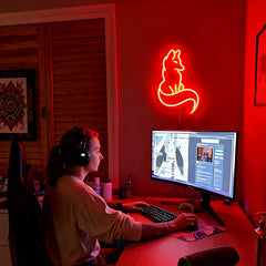 Person sitting at a desk with headphones on, using a computer in a room with a neon fox sign on the wall.