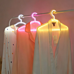 Three silky garments hanging on illuminated neon hangers in white, hot pink, and lemon yellow light, with one patterned with small multicolored hearts, displayed on a clothing rack against a neutral background.