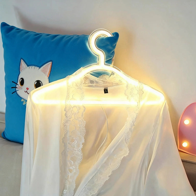 White lace-trimmed garment hanging on a glowing neon hanger, displayed in front of a blue pillow with a cartoon cat face, with lamp in the background.