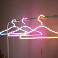 Three glowing neon clothing hangers in white, hot pink, and lemon yellow, hanging on a clothing rack against a neutral background.