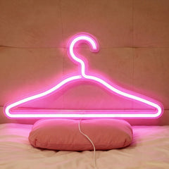 Bright hot pink neon clothing hanger light resting on a pink pillow against a beige headboard, with a power cord visible at the bottom.