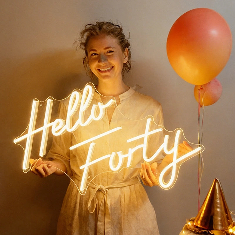 A smiling woman in a light warm white robe holds a glowing neon sign that reads 'Hello Forty' against a wall. Two orange balloons and a gold party hat are visible beside her.