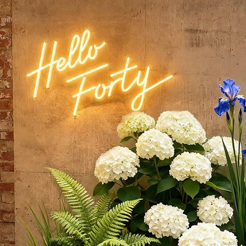 A glowing warm white neon sign that reads 'Hello Forty' mounted on a textured beige wall, with white hydrangeas, green ferns, and a blue iris in the foreground.